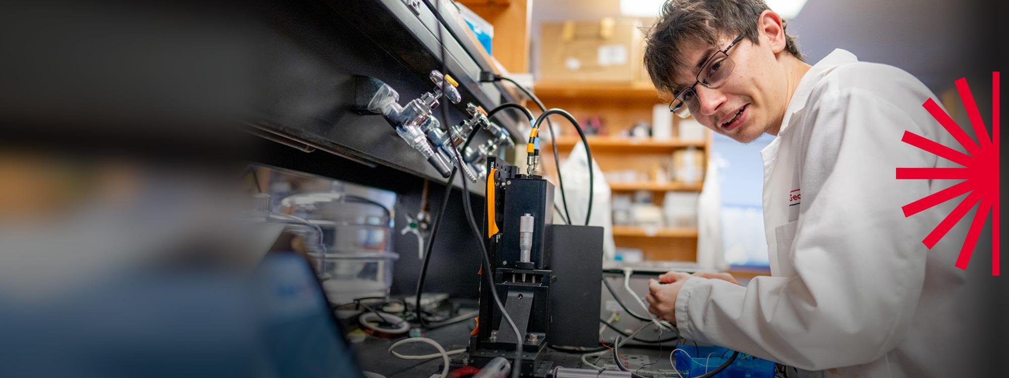 An undergraduate student is wearing a white coat and working in the Physics lab